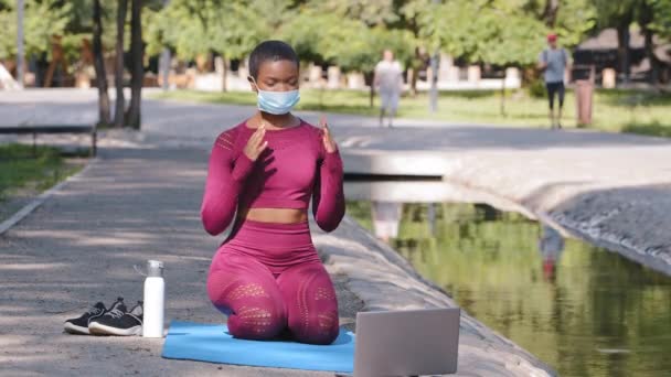 Instructora afroamericana americana que imparte clases en línea, practicando yoga meditando en la esterilla durante la cuarentena. Jovencita en ropa deportiva y máscara médica protectora con portátil sentado al aire libre — Vídeo de stock