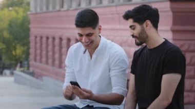 Bir çift çekici Hintli genç Arap açık havada oturuyor, cep telefonlarını tutuyor, akıllı telefon ekranlarına bakıyor, tatille ilgili izlenimlerini paylaşıyor, şov fotoğrafları çekiyor, hafta sonunun tadını çıkarıyorlar.