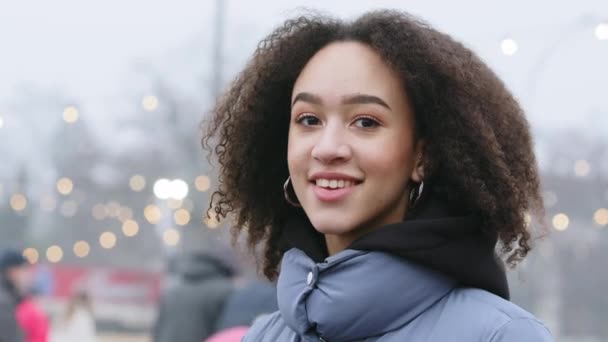 Side view of female portrait of African American girl with stylish afro hairstyle in curls stands outdoors on background of city skating rink and people, brunette looks at camera and smiles toothy — Stock Video