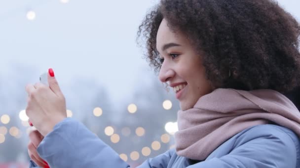 Side view young attractive afro american woman with curly hair in warm outerwear standing at city park in winter time, looking at her phone online dating chat in app social network smiling smis — Wideo stockowe