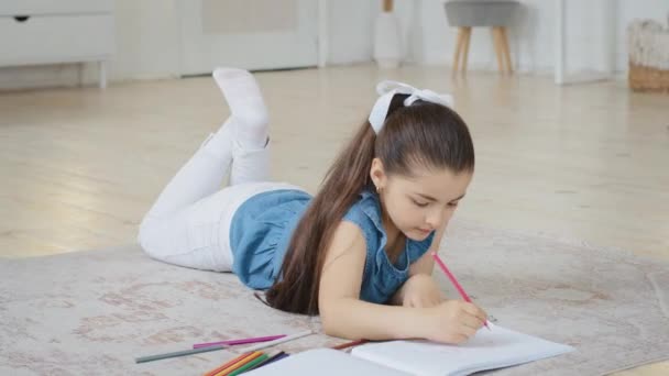 Spanish spaniard caucasian little small school girl daughter child pupil kid lying on floor draws picture drawing on paper painting with crayons enjoying hobby doing homework at home in living room — Stock Video