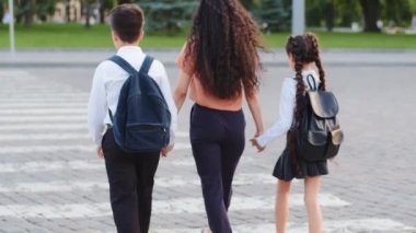 Ağır çekimde, iki çocuklu anne kadın iki öğrenci kız ve oğullarının ellerini tutuyor küçük kız ve küçük oğlan yaya geçidine basıyor şehirde yol boyunca yürüyor.