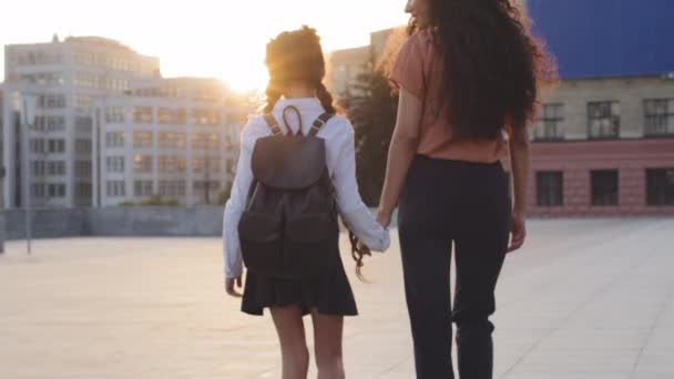 Indietro vista donna madre camminando lungo la strada alla luce del sole del tramonto con la bambina figlia studentessa con lo zaino che va da scuola. Mamma con bambino bambino cammina tiene femminile per mano rallentatore — Video Stock