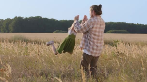 Opiekuńczy ojciec brodaty mężczyzna tata trzyma córeczkę dziecko dziecko dziecko przez ramiona wiry ręce obraca się w powietrzu maluch wirujący aktywnie gra stojąc na polu pszenicy na świeżym powietrzu letni dzień — Wideo stockowe