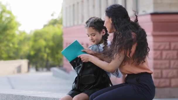 İspanyol anne, anne, anne, baba, sevgili kızıyla birlikte dışarıda oturuyor. Okul hazırlıklarına yardım ediyor. Okul çantasına not defteri koyuyor. — Stok video