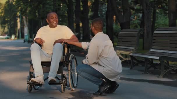 El joven negro le dice a su padre paralizado buenas noticias, habla de su éxito. Hombre afroamericano discapacitado en silla de ruedas se divierte comunicándose con su amigo o hermano feliz de conocer el resultado del caso — Vídeo de stock