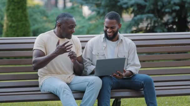 Δύο Αφροαμερικανοί φίλοι ή συνάδελφοι που κάθονται στον πάγκο στο πάρκο χρησιμοποιώντας φορητό υπολογιστή. Μαύροι άντρες που κοιτάζουν την οθόνη του υπολογιστή, λαμβάνουν μεγάλα email, συζητούν τα αποτελέσματα της συμφωνίας, χαίρονται τα καλά νέα — Αρχείο Βίντεο