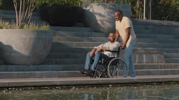 Afrikalı Amerikalı gülümseyen sakallı genç adam hafta sonları temiz hava alıp eğlenirken arkadaşı ya da gardiyanı güneşli bir yaz gününde şehir sokaklarında tekerlekli sandalyeyi itiyor. — Stok video