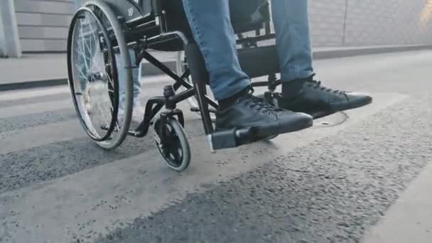 特写。男性亲属、护理人员或医务工作者帮助无行动能力、瘫痪、无行走能力的病人过马路。现代轮椅的轮子缓缓地从车行道升到人行道上 — 图库视频影像