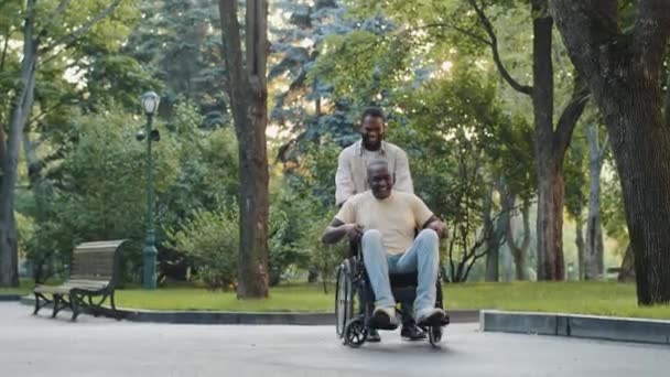 Cheerful young black guy visits brother in hospital post-op, walks, has fun in park with relative or friend. African man pushes father paralyzed after injury, surgery, disabled person in wheelchair — Stock Video