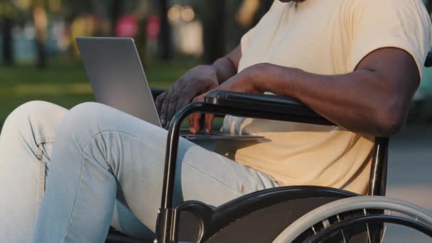 Die Nahaufnahme eines erwachsenen, gelähmten Afroamerikaners, der Text auf der Computertastatur tippt, kommuniziert in sozialen Netzwerken mit E-Mails. Behinderte im Rollstuhl arbeiten mit Laptop im Freien — Stockvideo