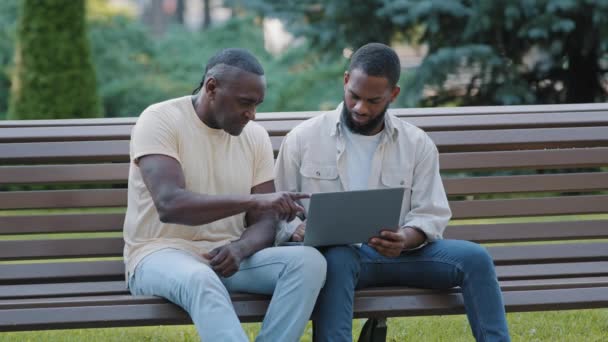 Двое черных мужчин в повседневной одежде сидят на скамейке в городском парке, используя ноутбук. Коллеги или друзья смотрят на экран компьютера, изучают новое приложение, просматривают фотографии, проверяют электронную почту на открытом воздухе — стоковое видео