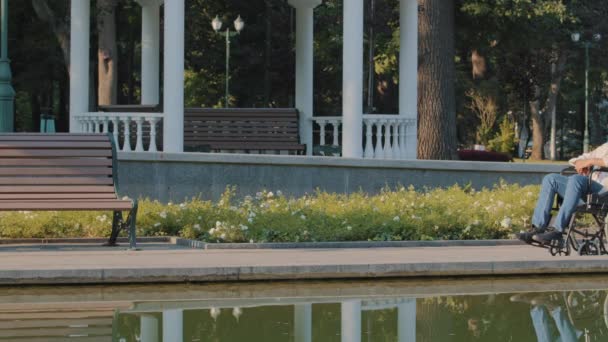 Joven hombre discapacitado disfruta de caminar en el parque de la ciudad por el agua en el soleado día de verano. El acompañante o tutor acompaña al paciente en silla de ruedas. El cuidado de las personas con discapacidad. Persona con movilidad limitada — Vídeos de Stock