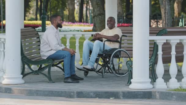 Gammal svart man och hans unga afrikanska manliga vän sitter utomhus och pratar om perspektiv, har roligt tillsammans på helgerna. Killen besöker sin far på sjukhus park under rehabilitering — Stockvideo