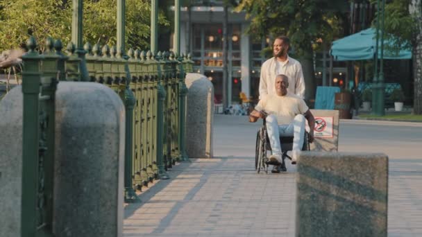 Afrikansk invalid man. Svart man handikappad. Gammal vuxen handikappad person i rullstol promenader med son eller vårdnadshavare längs stads gränd på solig sommardag, njuta av vila, ha god tid på helgen — Stockvideo