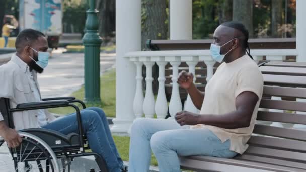 Deux Afro-Américains portant des masques médicaux se retrouvent à l'extérieur, se saluent avec des coudes, respectent les règles de quarantaine. Noir jeune handicapé en fauteuil roulant accueille ami, discuter des dernières nouvelles — Video