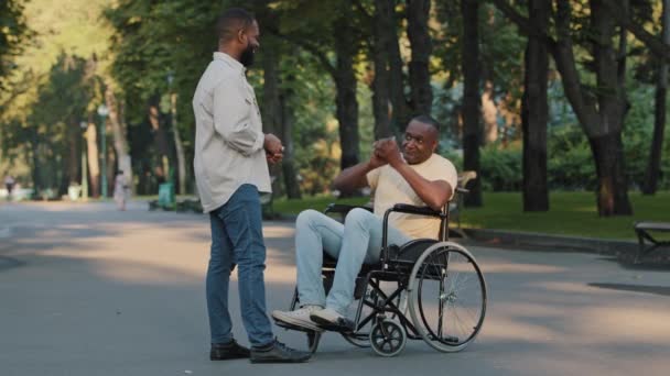 Dos hombres africanos relajándose en el parque del hospital, divirtiéndose al aire libre, charlando hablando del campeonato. Negro hombre discapacitado en silla de ruedas gesticulando vigorosamente mientras discute juego de baloncesto con un amigo — Vídeos de Stock