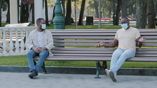 Two African American men in facemask sitting on bench, keeping distance, discussing problems of teleworking during quarantine. Black male friends talk about wash and disinfect hands due to epidemic — Stock Video