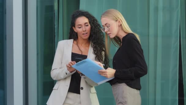 Millennial European Caucasian woman executive ceo discuss report with young Indian colleague, serious multiracial female employees read paper discussing business plan talk outdoor near building — Vídeo de Stock