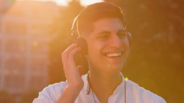 Alegre joven milenario de Oriente Medio divertirse disfrutando de pistas en los auriculares, emocionado sonriente indio árabe macho escuchar música en los auriculares al aire libre noche de verano, luz del sol brillante luz de fondo — Vídeos de Stock