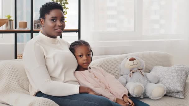 Afro americká rodina usměvavá mladá matka žena chůva maminka a vážné holčička dítě dítě dítě sedí mazlení na pohovce doma v obývacím pokoji při pohledu na kameru, černá adopce koncept — Stock video