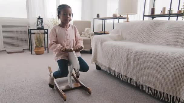 Little afro amerykanka dziewczyna czarny dziecko małe dziecko uczennica córka siedzi na drewnianym koniu huśtawka na krzesło zabawka gra w domu w salonie ciesząc się grą udając jeźdźca jeźdźca — Wideo stockowe