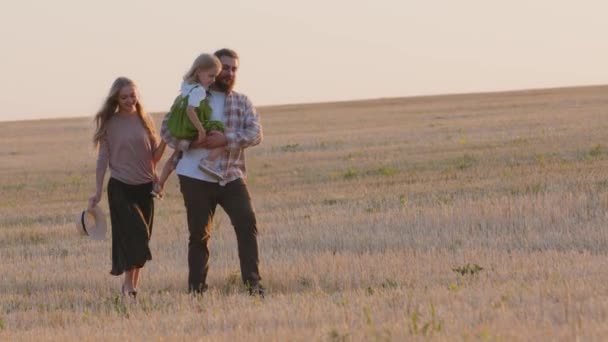 Caucasian family farmers parents with small daughter walking along wheat field in rural landscape. Mother woman and man father holding in arms baby girl going walk outdoors on yellow grass talking — Stock Video