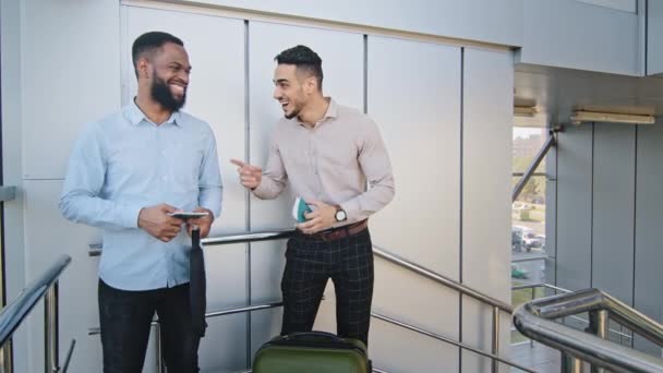 Two business men multiracial colleagues multiethnic co-workers hispanic man and afro american guy standing in terminal airport talking laughing looking at camera holding boarding passes plane tickets — Stock Video