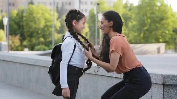 Родительская мать брюнетка выпрямляет косички дочери говорящей ребенок говорит улыбаясь школьнице ученик перед классами обнимает обнимает латиноамериканка девочка обнимает маму на открытом воздухе обратно в школу — стоковое видео
