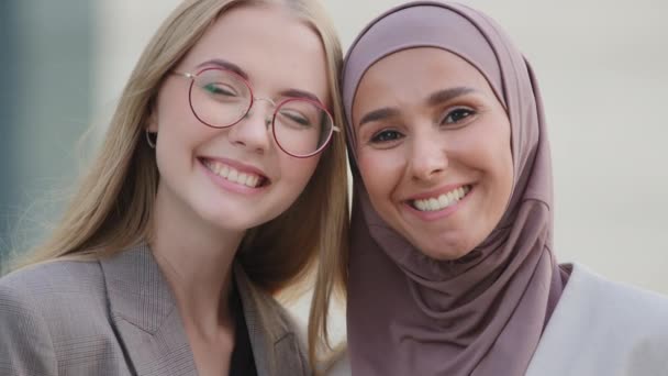 Felices colegas o novias del milenio se acurrucan contentos de conocerse. Muchacha india sonriente en hijab y joven europea con gafas de pie juntas, amistad femenina, solidaridad — Vídeo de stock