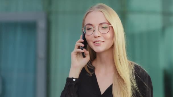 Happy young woman in glasses caller talking on phone outdoor, cheerful millennia girl enjoys pleasant mobile conversation, smiling Caucasian female holding cell speaking making call by telephone — Stock Video