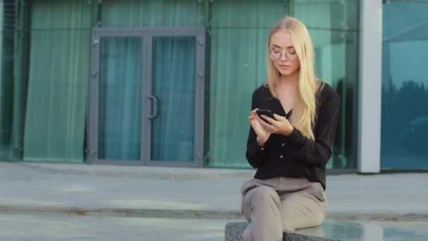 Blonde jeune étudiante caucasienne assise en plein air tenant téléphone mobile surf internet a reçu un message de lecture. Une fille européenne salue un collègue indien. Femme orientale rencontre avocat ou employeur — Video