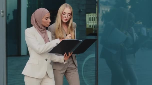 Deux femmes d'affaires diverses marchant le long de la rue tenant dossier avec des documents communiquent avant la réunion, discuter rapport. Indienne en hijab consulte une collègue jeune femme européenne pendant la pause — Video