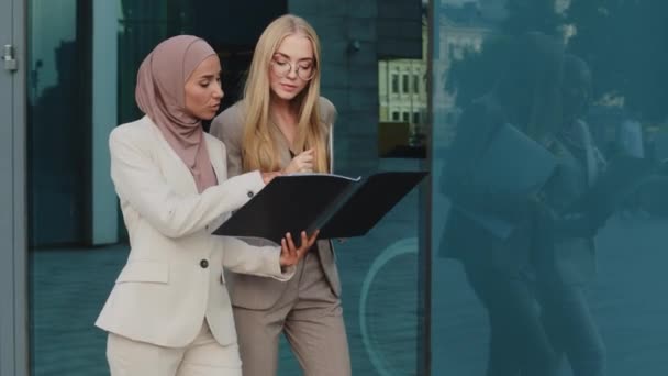 Dos empresarias diversas caminando por la calle sosteniendo carpeta con documentos se comunican antes de reunirse, discutir informe. India chica en hijab consulta con colega joven europea durante el descanso — Vídeos de Stock