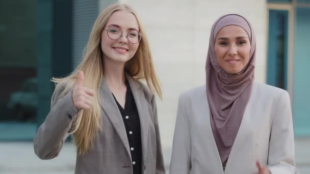 Zwei fröhlich lächelnde junge Wirtschaftsführerinnen, die Daumen hoch machen. Kopfschuss Porträt vielfältiges Team, multiethnische Gruppe. Zufriedene indische Kundin im Hijab, Europäerin gibt positives Feedback — Stockvideo