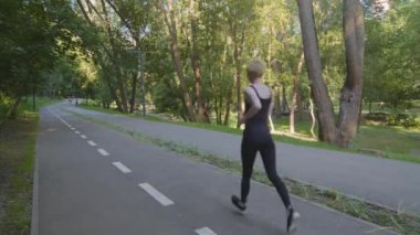 Beyaz tenli, ince, güçlü, motive olmuş kadın koşucu, kısa saçlı, siyah spor giyimli koşucu. Koşu parktaki koşu pistinde koşuyor.