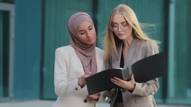 Две разноплановые коллеги-деловые женщины встречаются, беседуют, просматривают документы. Индийская девушка в хиджабе и молодая европейская женщина, работающая вместе, неформально общаются во время перерыва на работу — стоковое видео