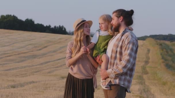 Familia de agricultores caucásicos se encuentra en el campo de trigo mira el paisaje de la naturaleza rural horizonte. Los padres jóvenes sostienen pequeña hija niña charla comunicación conversación disfrutar de la vista apuntando dirección con la mano — Vídeos de Stock