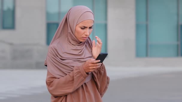 Muslim islamic girl woman in hijab stands on street outdoors city uses mobile phone looks at smartphone screen cellphone reads bad news online feels sad disappointment receives refusal notification — Stock Video