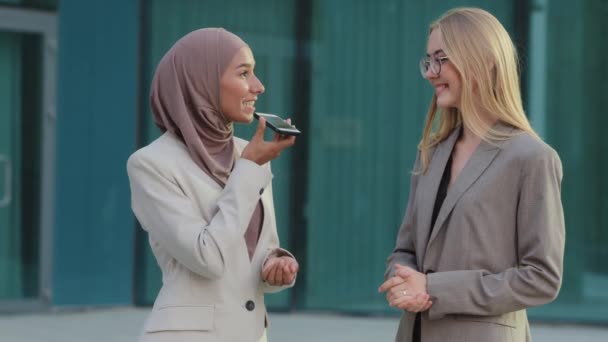 Zwei verschiedene professionelle Geschäftsfrauen Kolleginnen verwenden mobile Anwendung, um Sprachproben oder Assistenten aufzunehmen. Millennial East Indian Girl im Hijab bittet junge blonde Frau, Wort per Telefon zu übersetzen