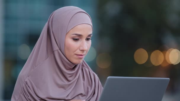 Yakın planda odaklanmış müslüman iş kadını internette dizüstü bilgisayarla tesettüre takılır, internette çalışır, kameralara bakar, el hareketi yapar. Şehirdeki bilgisayarı olan İslami kız işaret veriyor. — Stok video