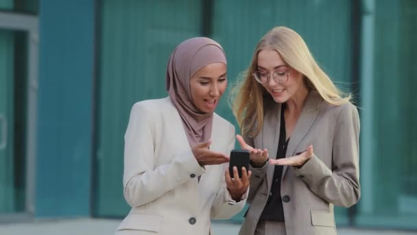 Glad varierande Unga kvinnliga kollegor vilar under kontorspausen, blond kaukasisk flicka och tusenårig orientalisk kvinna i hijab håller mobiltelefon surfa internet titta på bilder eller roliga videor — Stockvideo