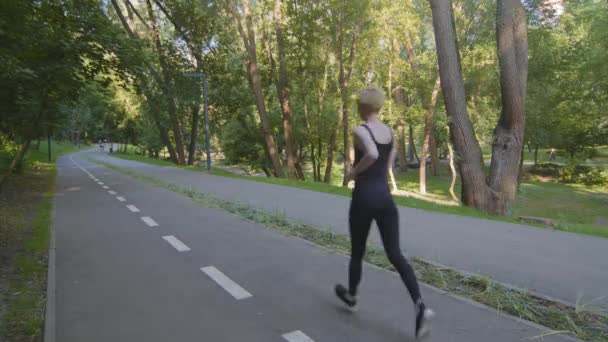 Вид сзади в полный рост кавказский стройный сильный активный мотивированный женщина блондинка бегун бегун с короткими волосами в черной спортивной одежде работает на беговой дорожке в парке утром бег трусцой спорта — стоковое видео