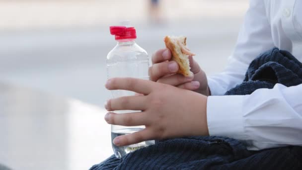 Cropped filming unrecognizable hungry child boy girl pupil kid in white shirt with backpack schoolbag holding bun food baking sweet snack and bottle of clear still water eating bites eat meal outdoors — Stock Video