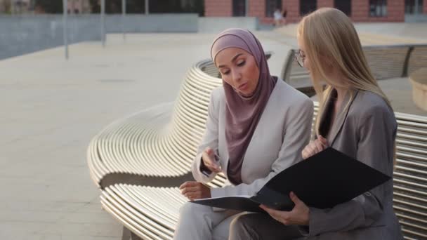 서류를 들고 폴더를 들고 사무실 문을 열고 있는 두 명의 다양 한 실무인 여성 이 야외에 앉아 대화를 나눈다. 히잡에 있는 인도 소녀가 동료 유럽 여자와 상담하는 모습 — 비디오