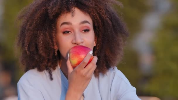 Afro-americana mulher africana encaracolado menina sentado ao ar livre farejadores vermelho fresco maduro maçã deliciosa fruta desfrutar de dieta saudável regozija-se cheiro retorno após coronavírus doença fala olhando para a câmera — Vídeo de Stock