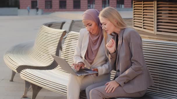 Smiling business women sitting outdoors with laptop looking at screen, satisfied result showing like thumb up. Diverse female students, hijab Indian girl and young European woman wearing glasses — Stock Video