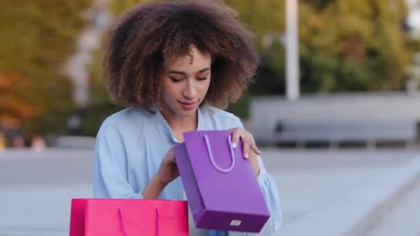Afroamerykanka Afro młoda afrykańska dziewczyna siedzi na ulicy na zewnątrz trzymając torby prezentowe patrzy w pakiecie zakupu zaskoczony raduje się niespodzianką prezent pokazuje w porządku ręka gest ok znak, koncepcja zakupów — Wideo stockowe