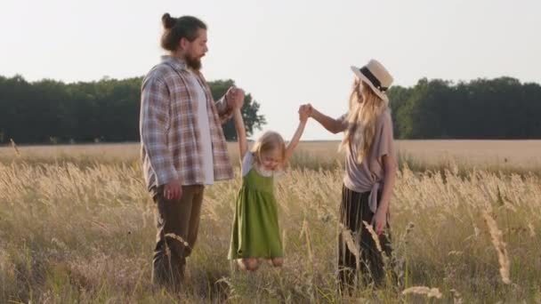 快乐的父母三个人带着孩子站在麦田里在大自然里男人和小孩在外面玩耍牵着女儿的手把小女儿抚养成空中秋千飞飞的游戏 — 图库视频影像