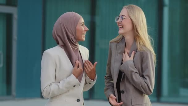 Ung Blond Kaukasiska flicka i kostym prata med vän, orientalisk tusenårig kvinna i hijab dela nyheter med kollega. Kvinnliga anställda vilar skvaller och skrattar under kontorspausen — Stockvideo